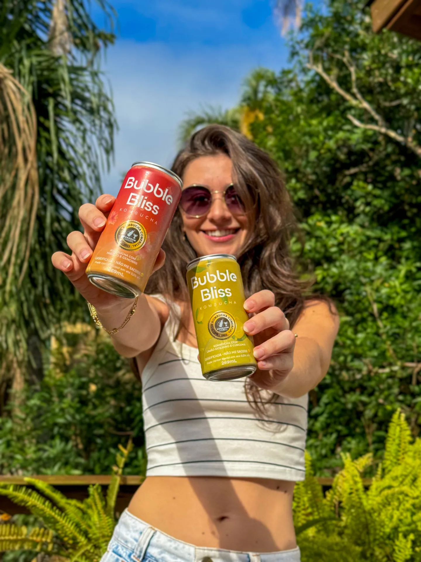 Cliente sorrindo no jardim segurando duas latas Bubble Bliss — hibisco e limão siciliano